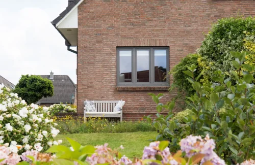 Anthrazitfarbene Aluminium-Fenster in Backsteinfassade mit blühendem Garten davor.
